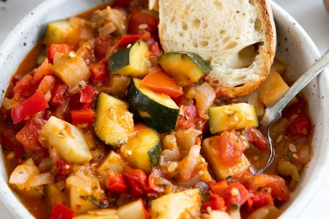 A bowl of vegetable stew with chunks of zucchini, tomato, and red pepper, topped with a slice of bread and a silver fork.