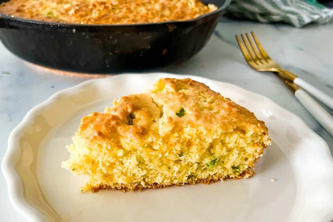 A slice of cornbread sits on a white plate in front of a pan of cornbread. Two forks are placed on the table in the background.
