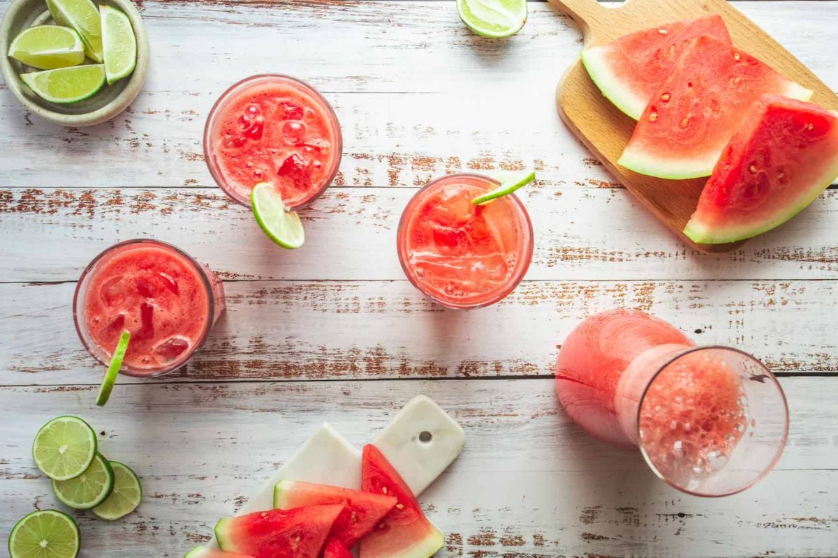 Top view of watermelon drinks with ice and lime slices in glasses, a pitcher, and watermelon wedges on cutting boards on a white wooden surface.