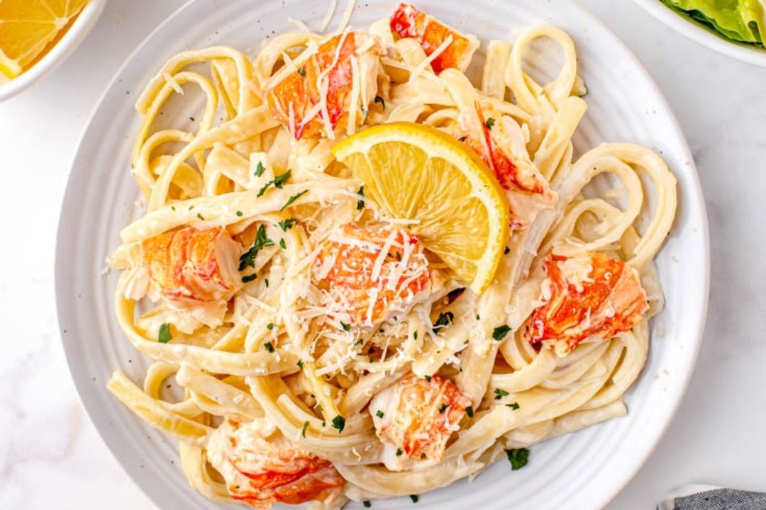 Fettuccine pasta with pieces of lobster, grated Parmesan cheese, parsley, and a lemon wedge on a white plate.