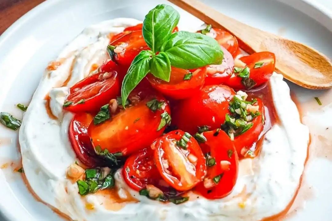 Cherry tomatoes with herbs on a bed of creamy yogurt, garnished with a basil leaf, served on a white plate with a wooden spoon.