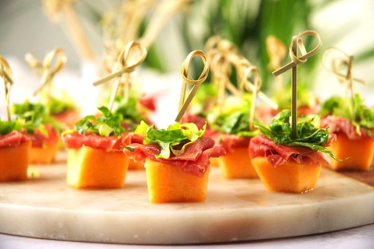 Bite-sized appetizers with prosciutto, greens, and melon cubes, each held together with a wooden skewer, arranged on a marble serving tray.