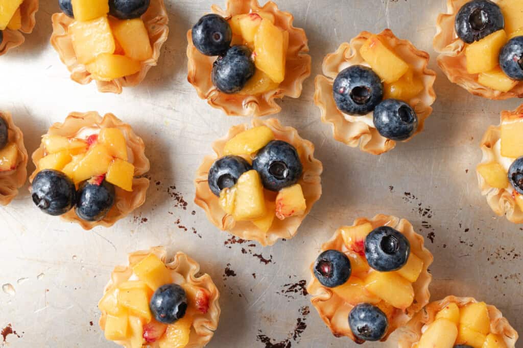 Mini tart shells filled with diced peaches and whole blueberries, arranged on a metal surface.