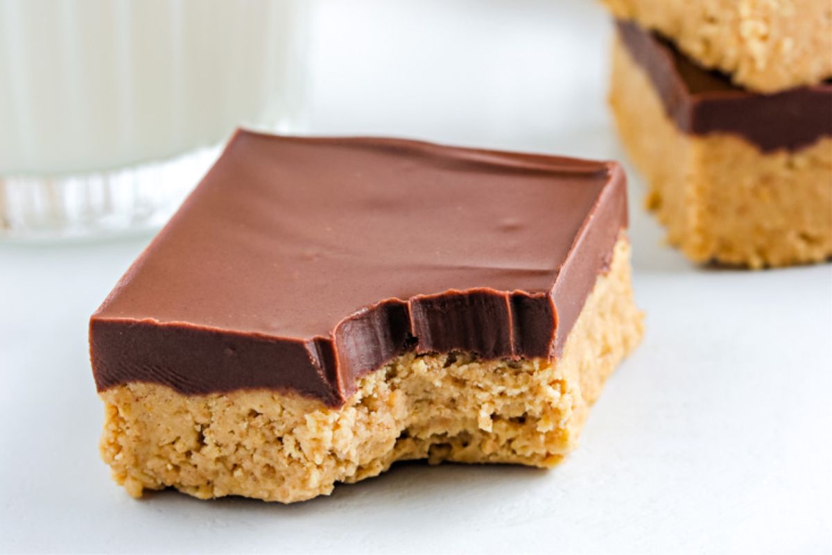 A peanut butter bar with a chocolate topping and a bite taken out, placed on a white surface next to a glass of milk.