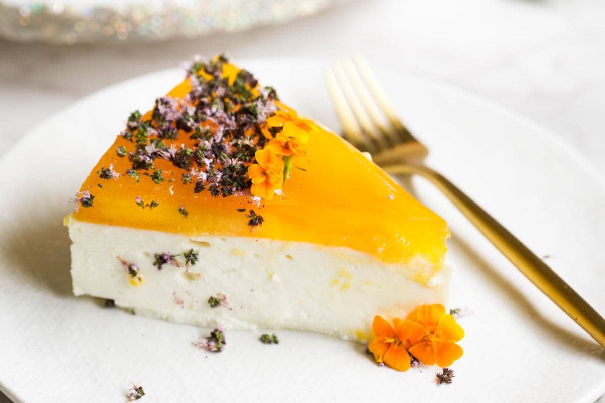 A slice of cheesecake with a yellow fruit glaze and edible flowers on top, served on a white plate with a gold fork.