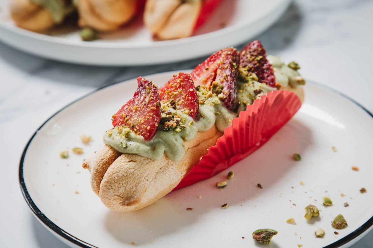 A dessert with a meringue base, topped with green pistachio cream, sliced strawberries, and crushed pistachios, served in a red paper liner on a white plate.