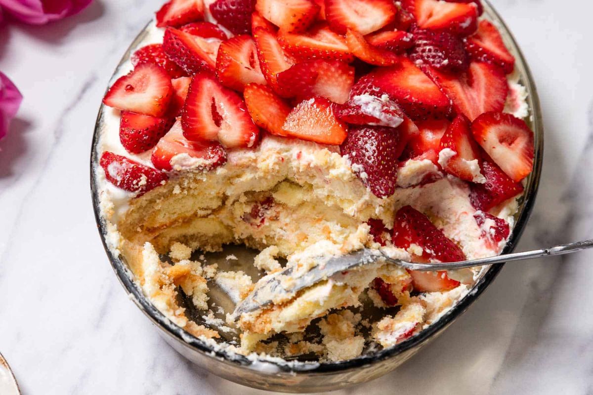 A glass dish of strawberry tiramisu with layers of cream and sliced strawberries, partially served with a spoon, on a white surface.