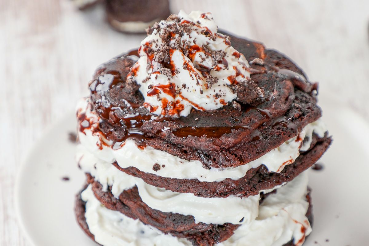 A stack of chocolate pancakes layered with whipped cream, topped with whipped cream, chocolate syrup, and cookie crumbles on a white plate.