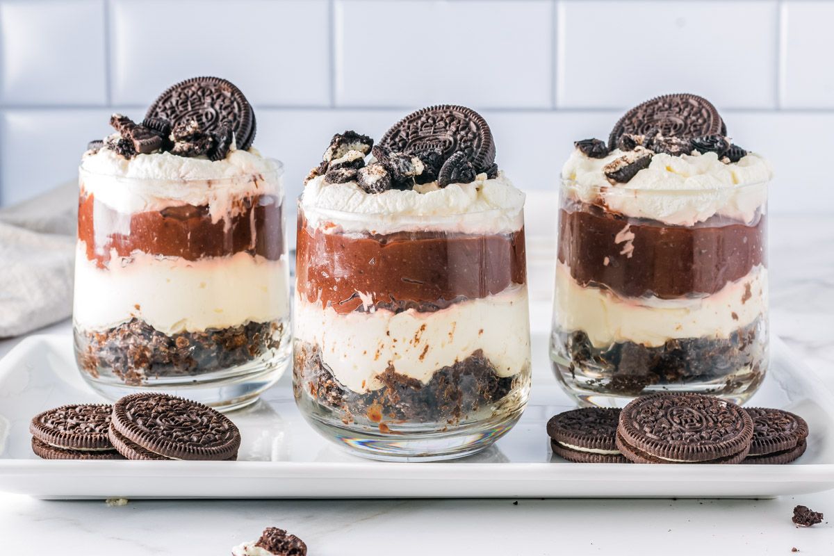 Three glass cups filled with layered Oreo dessert, including crushed cookies, cream, and chocolate pudding, topped with whipped cream and an Oreo cookie, displayed on a white tray.