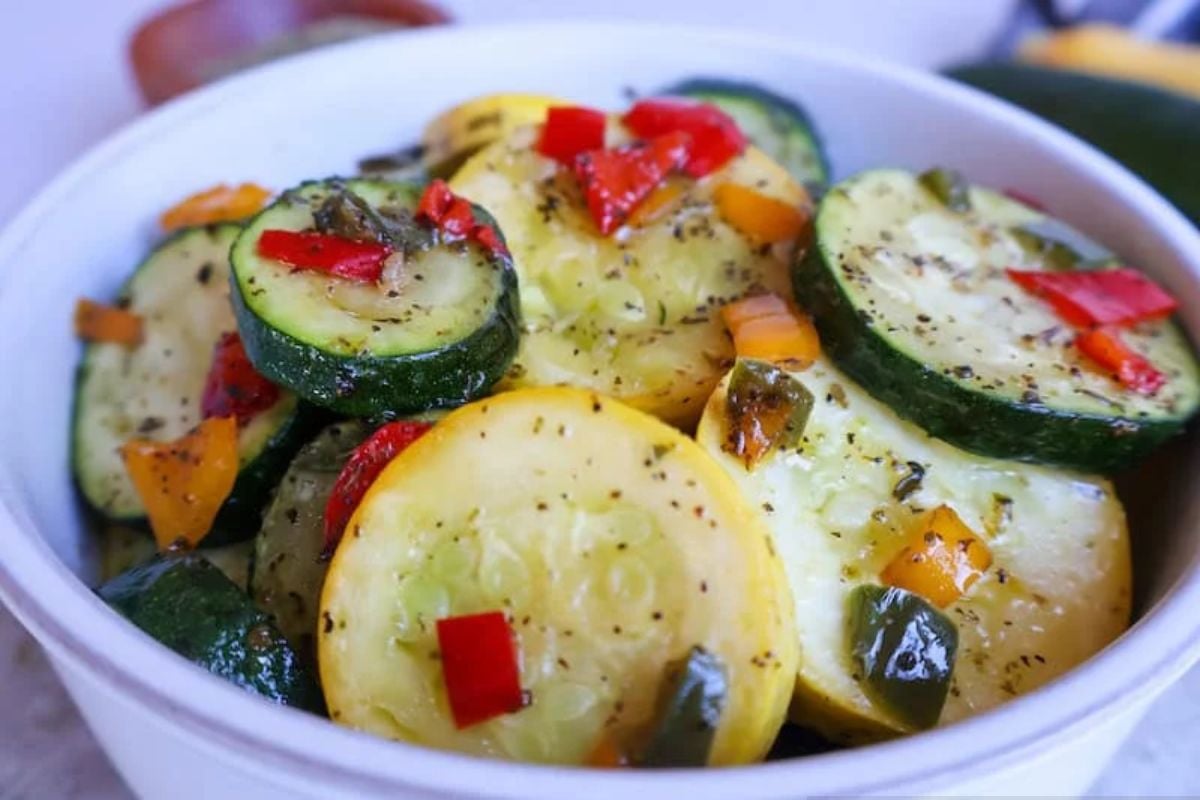 A white bowl filled with sliced zucchini and yellow squash, mixed with red and green bell peppers, topped with herbs and seasonings.