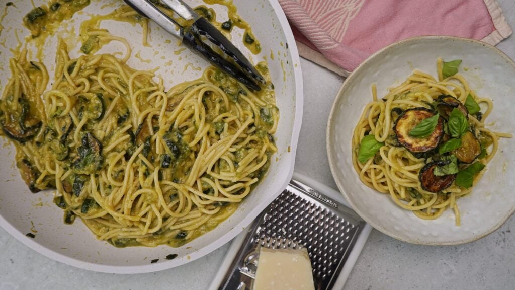 A pan of creamy spaghetti with vegetables next to a bowl of the pasta, fresh basil, a cheese grater, and a block of cheese on a white surface.