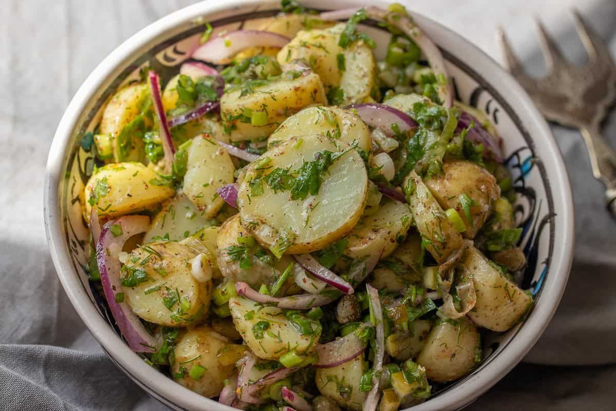 A bowl of potato salad with sliced red onions, chopped herbs, and seasonings, placed on a patterned dish beside a fork.