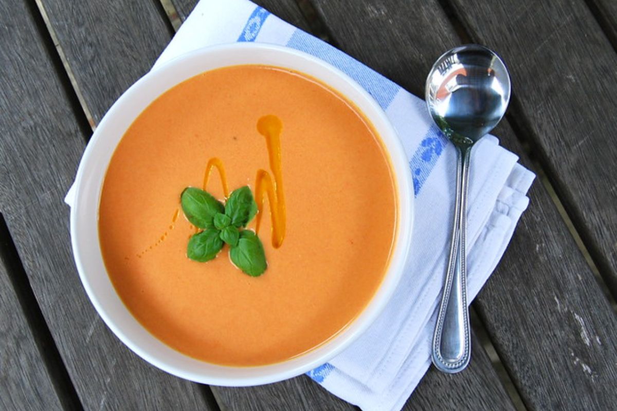 A bowl of creamy orange soup garnished with basil and a drizzle of oil, placed on a napkin with a spoon on a wooden table.