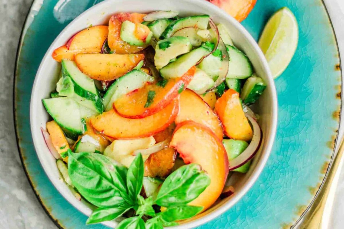 A bowl of salad with sliced peaches, cucumbers, red onions, and herbs, garnished with a basil sprig, on a blue plate with a lime wedge.