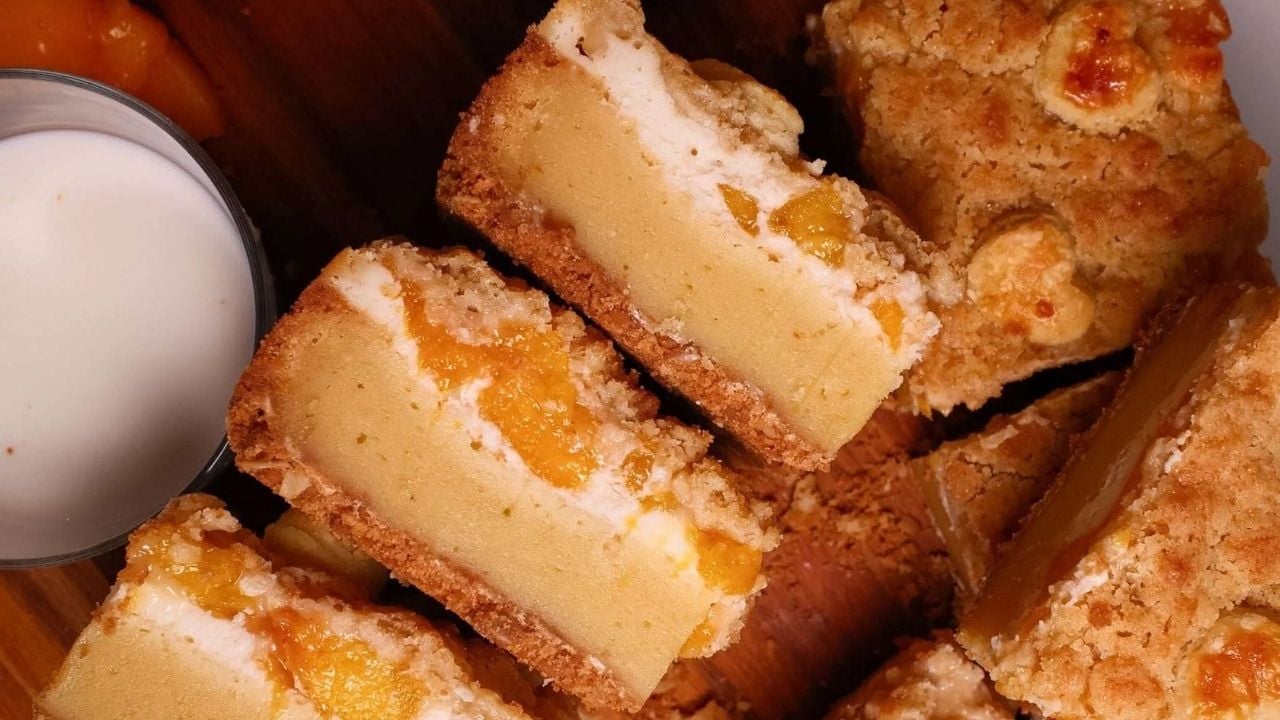 Sliced cheesecake bars with a crumbly crust and peach filling are arranged on a wooden surface next to a glass of milk.