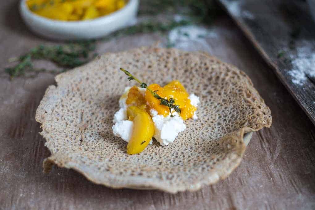 A piece of injera topped with white cheese, mango slices, and a sprig of fresh thyme on a wooden surface.