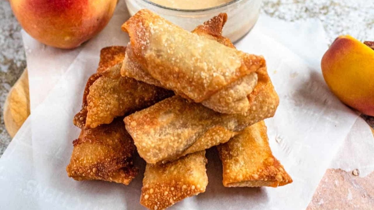 A stack of golden-brown fried egg rolls is displayed on parchment paper, with peach slices and a dipping sauce nearby.