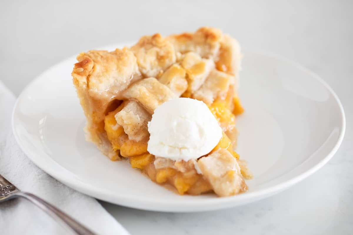 A slice of peach pie with a lattice crust sits on a white plate, topped with a scoop of vanilla ice cream.