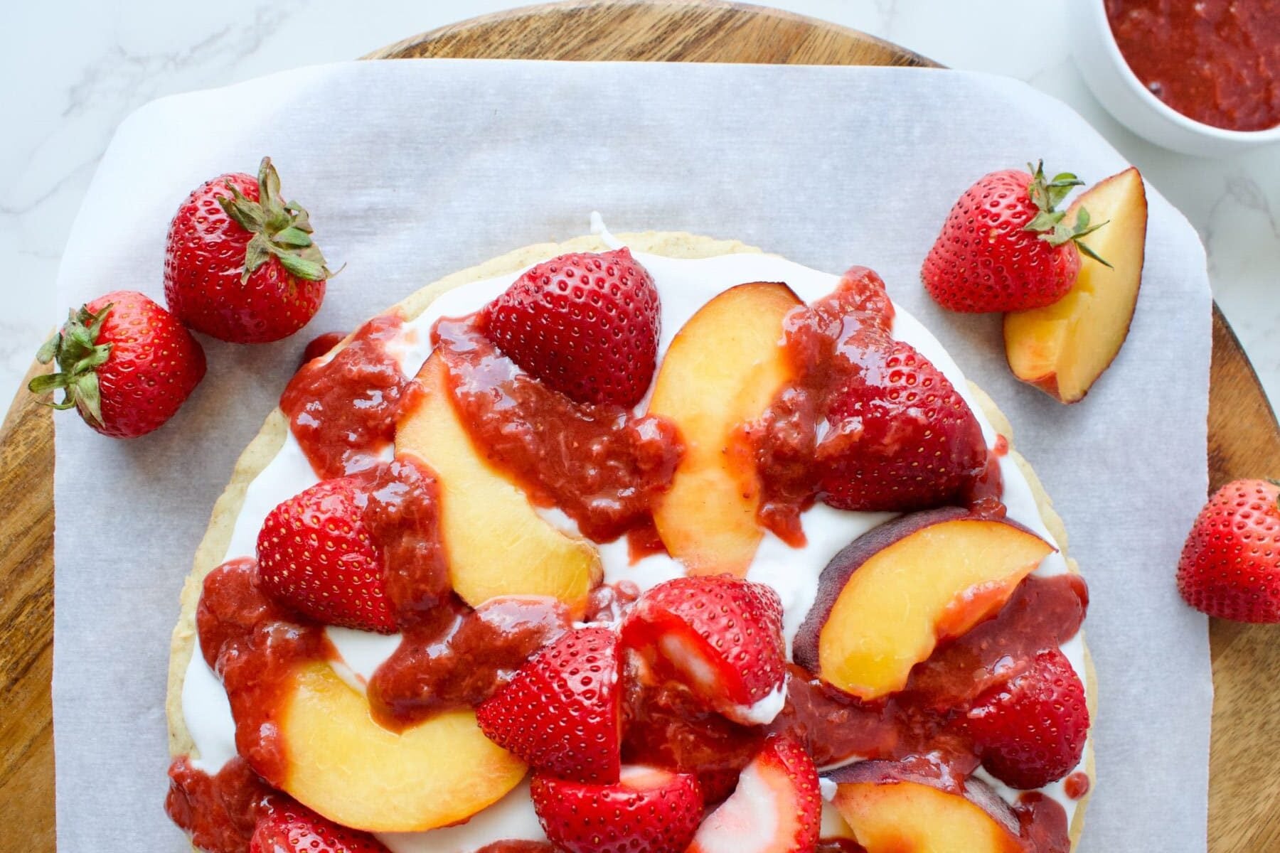 A dessert pizza topped with sliced strawberries, peach slices, strawberry sauce, and a creamy base, placed on parchment paper with whole strawberries around.
