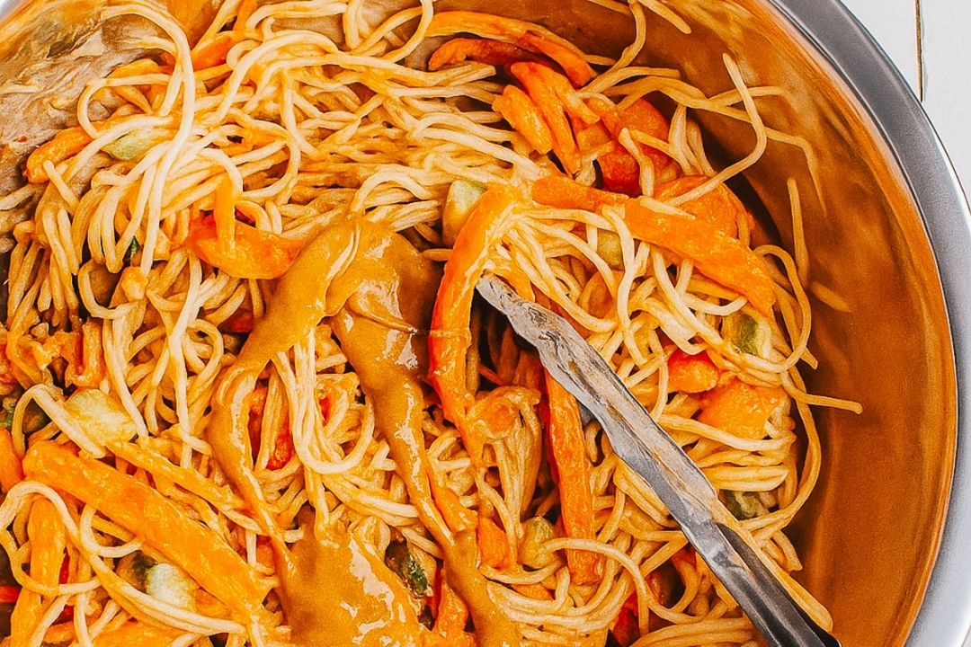 A bowl of noodles mixed with sliced vegetables and topped with a creamy brown sauce, with metal tongs resting inside.