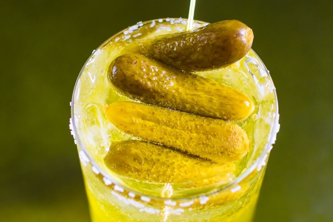 A close-up of a yellow-green drink with ice, garnished with several whole pickles on top, and a salted rim.