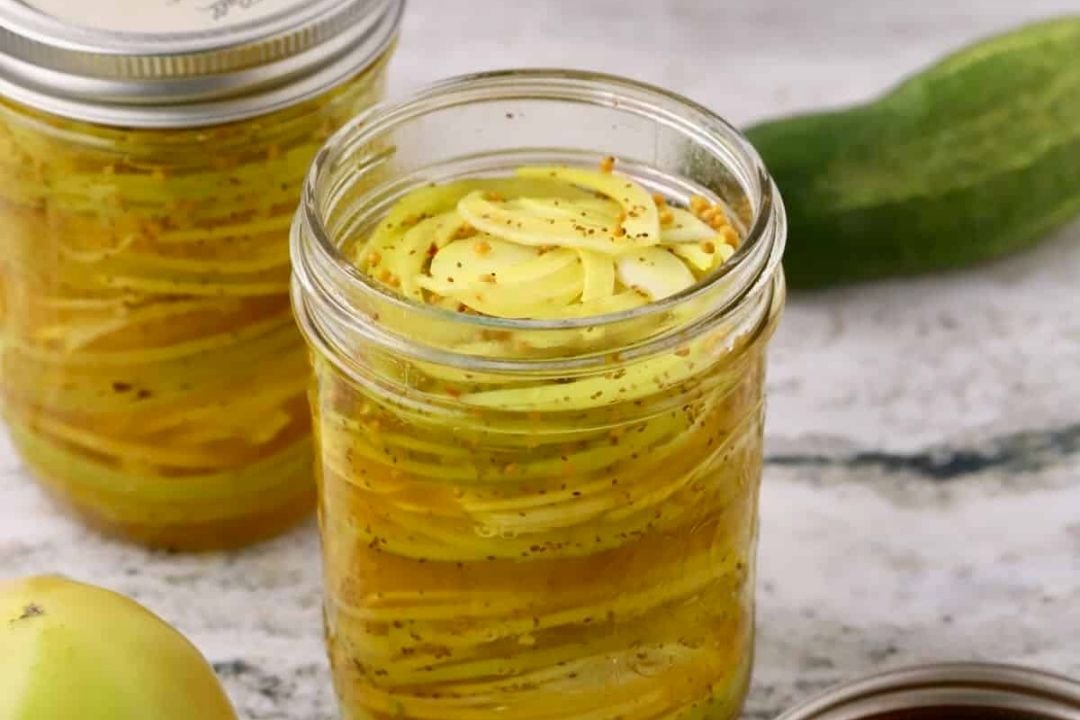 A glass jar filled with sliced yellow vegetables and spices in brine sits on a marble surface, with a cucumber and a second jar in the background.