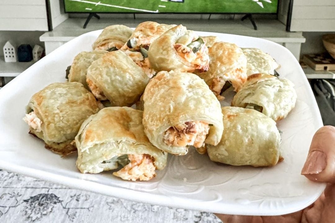 A hand holds a white plate of golden-brown puff pastry rolls filled with a creamy mixture, with a TV showing a sports game in the background.