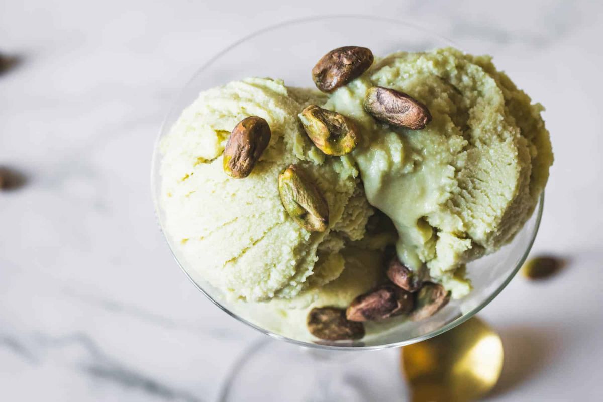 Scoops of pistachio ice cream in a glass dish, topped with whole pistachio nuts, on a marble surface.