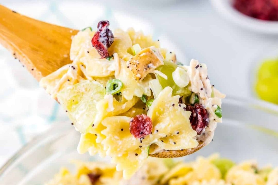 A wooden spoon holds bowtie pasta salad mixed with green grapes, cranberries, green onions, and poppy seeds.
