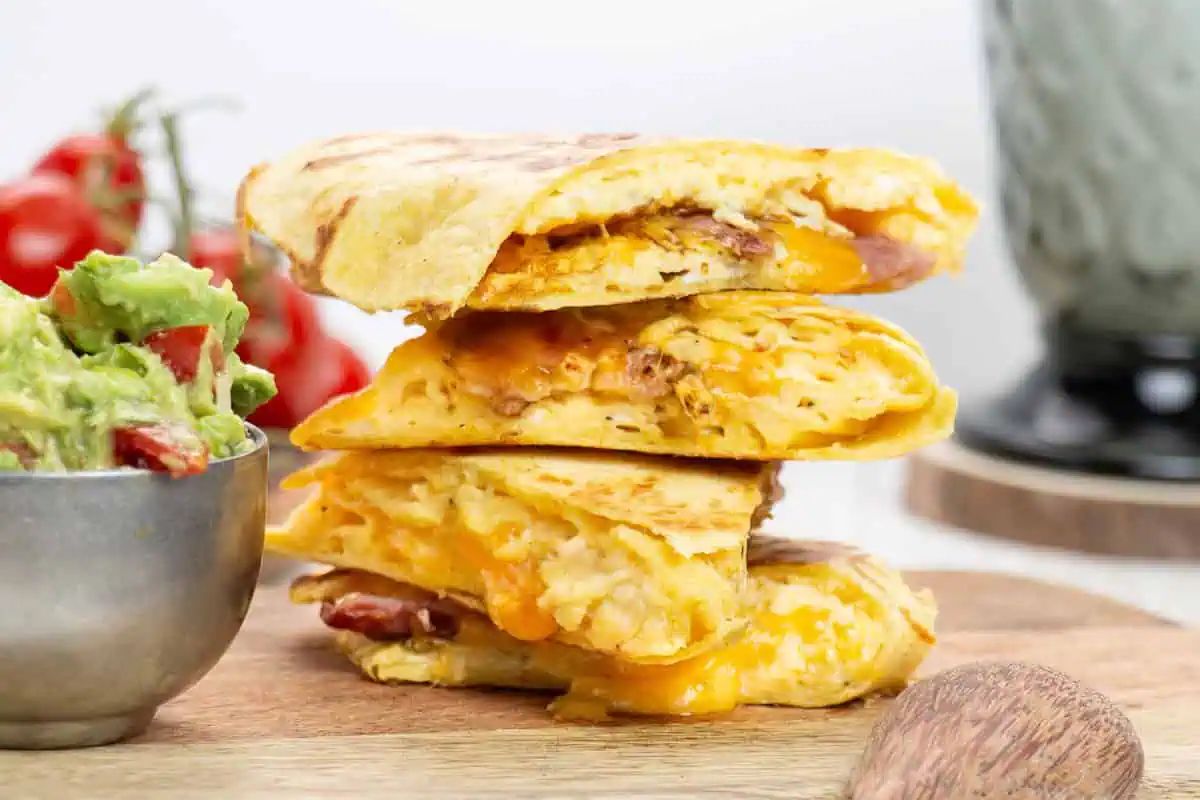 Three cheesy quesadilla wedges stacked on a wooden board, next to a bowl of guacamole and some tomatoes in the background.