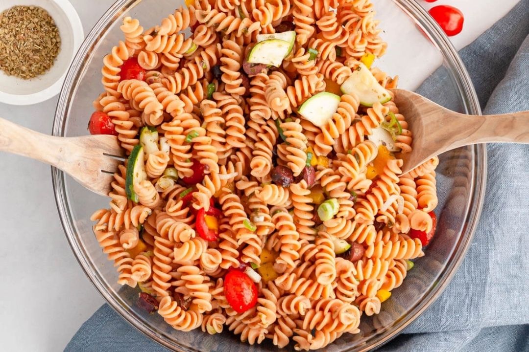 A glass bowl filled with rotini pasta salad containing chopped vegetables like tomatoes, cucumbers, and bell peppers, with two wooden serving utensils.