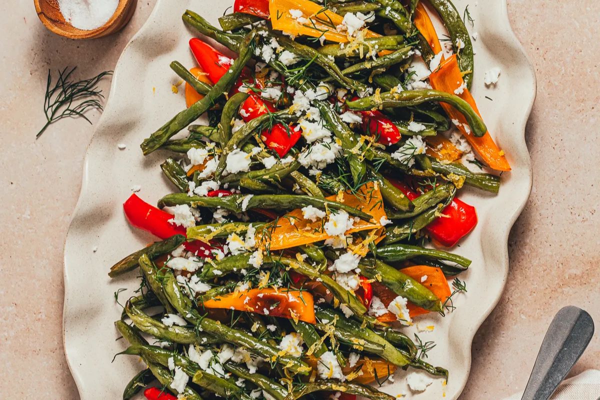 Roasted green beans and bell peppers topped with crumbled cheese, fresh herbs, and lemon zest are served on a wavy white platter. A small bowl of salt is visible in the background.