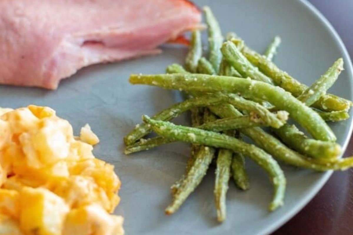 A plate with green beans, a slice of ham, and a serving of scrambled eggs or cheesy potatoes.