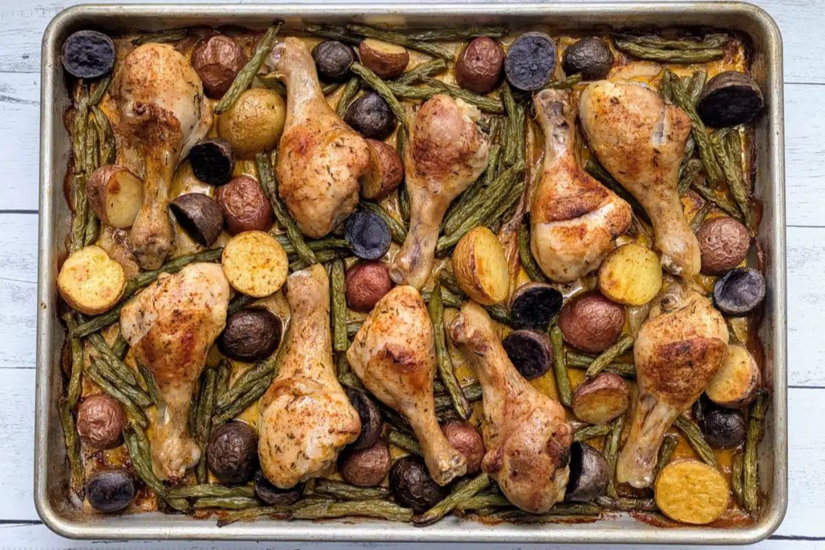 A sheet pan with baked chicken drumsticks, assorted halved potatoes, and green beans on a light wooden surface.