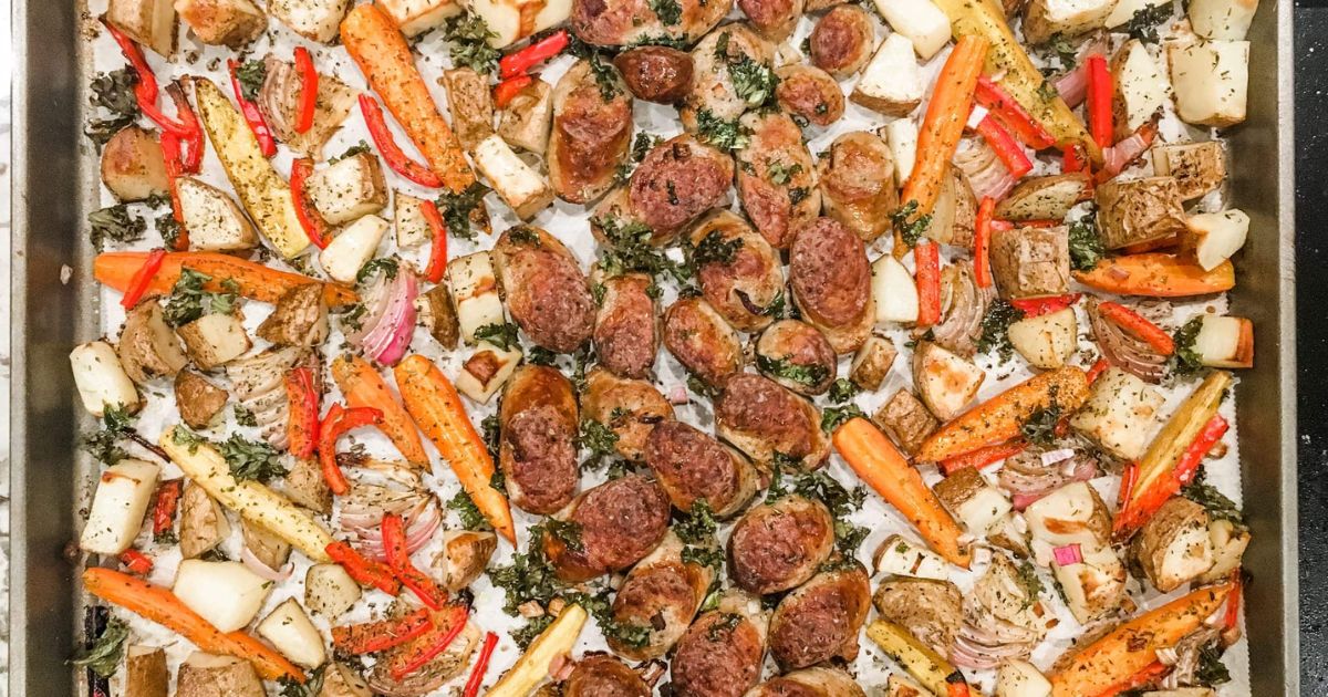 Sheet pan filled with roasted sausage slices, diced potatoes, carrots, parsnips, red bell peppers, onions, and herbs.