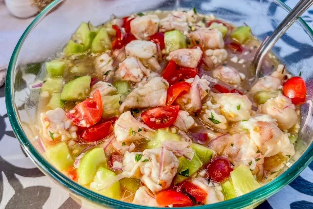 A glass bowl of shrimp ceviche with chopped cucumbers, tomatoes, onions, and herbs in a light marinade, with a spoon inside.