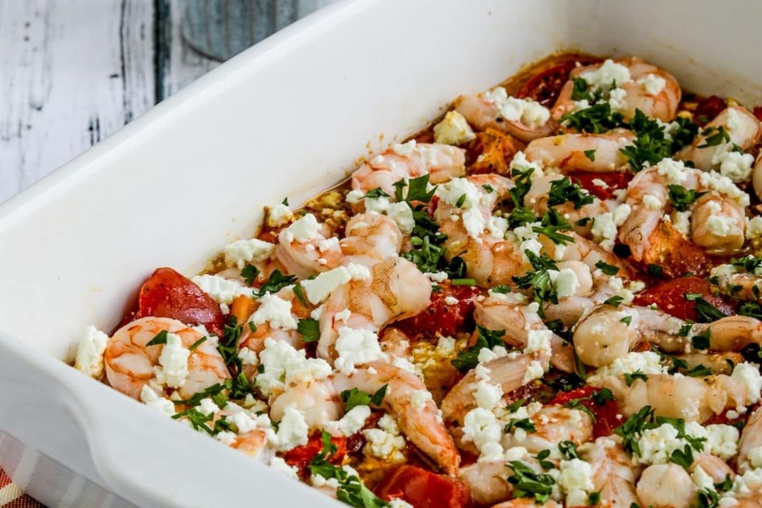 Baked shrimp with tomatoes, crumbled feta cheese, and fresh herbs in a white casserole dish on a wooden surface.