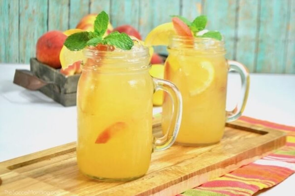 Two mason jars filled with iced peach lemonade, garnished with lemon slices and mint, placed on a wooden tray with peaches in the background.