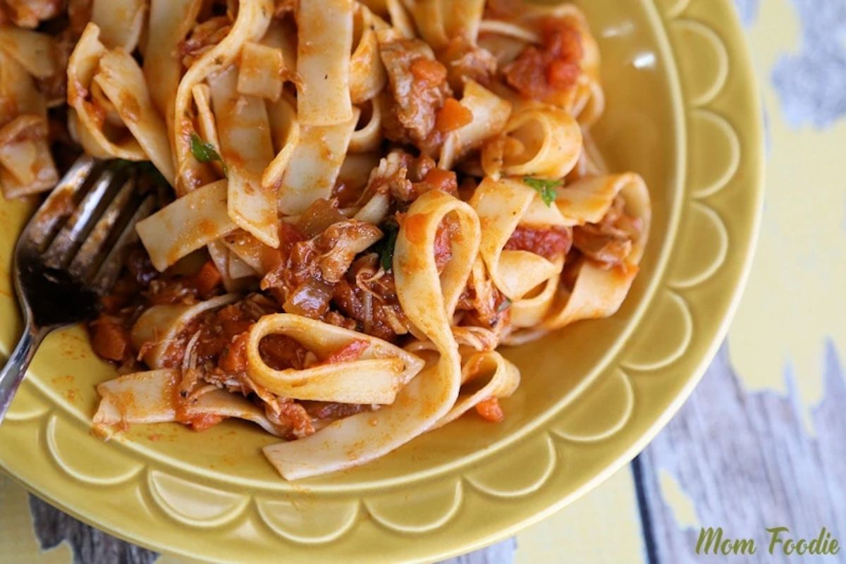 Wide pasta noodles with a chunky tomato-based sauce served on a yellow plate with a fork.
