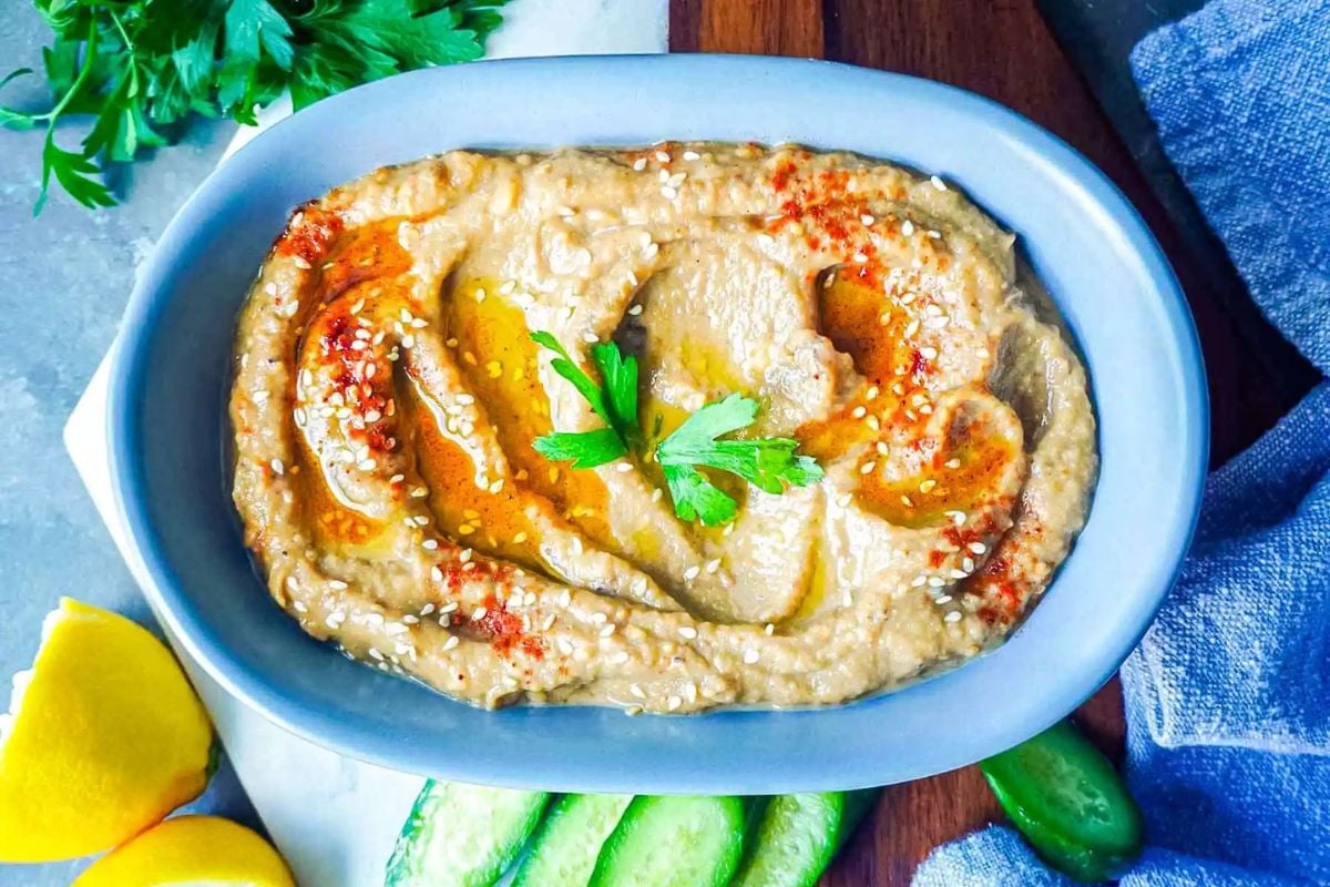 A bowl of baba ganoush garnished with olive oil, parsley, sesame seeds, and paprika, with lemon wedges, cucumber slices, and parsley on the side.