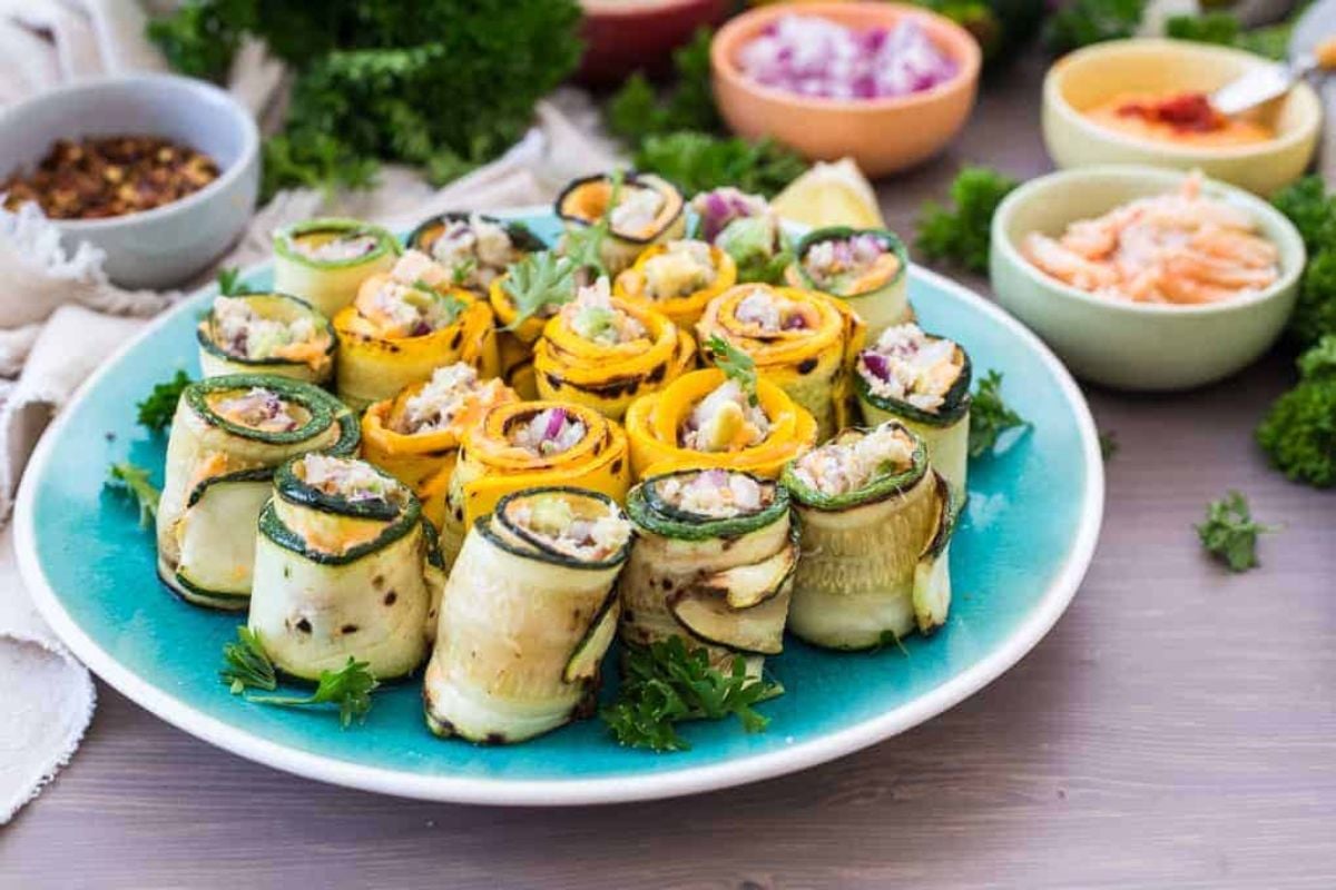 A plate of rolled zucchini and yellow squash appetizers filled with a creamy mixture, surrounded by small bowls of toppings and garnishes.