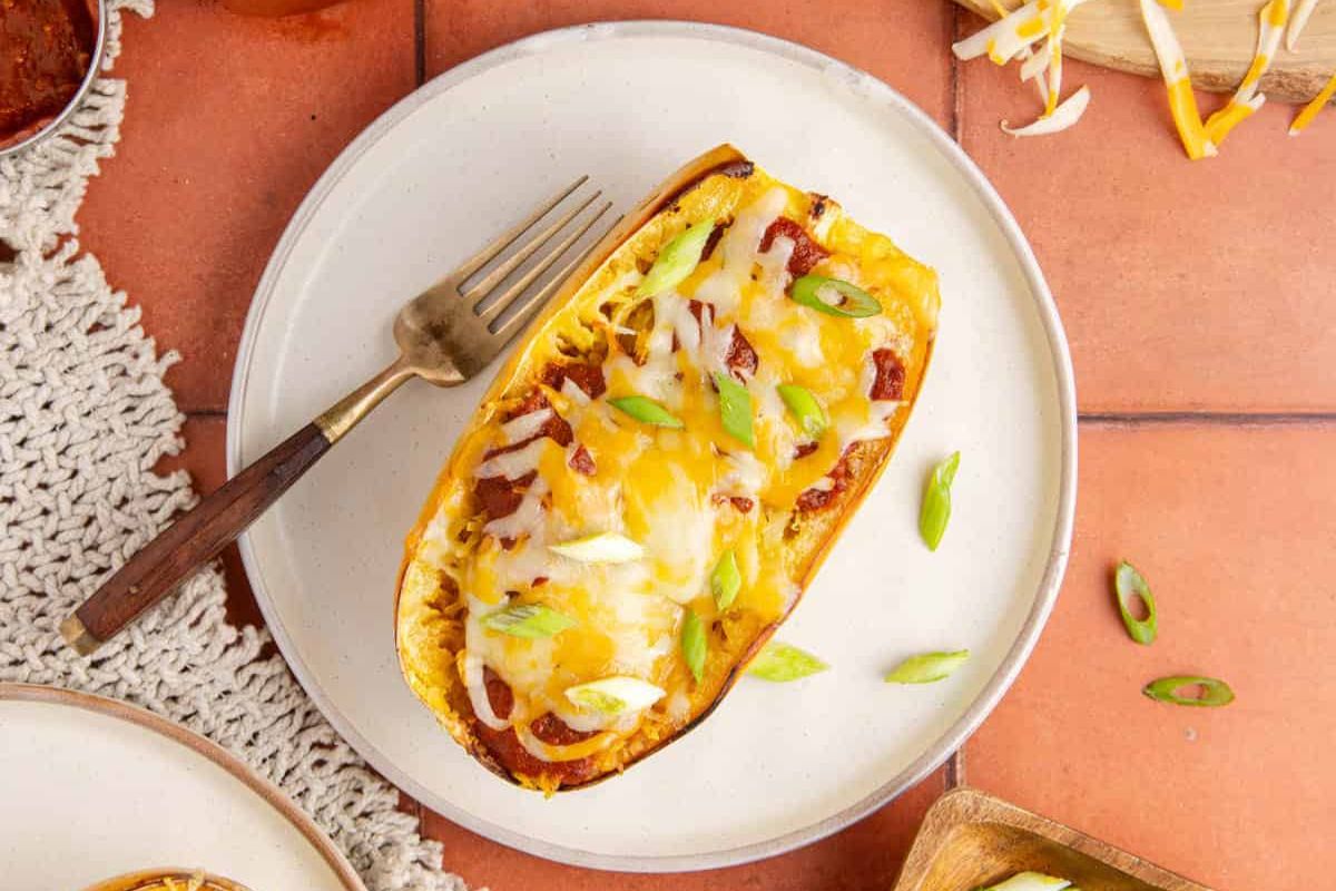 A halved spaghetti squash filled with cheese, sauce, and green onions on a white plate with a fork, placed on a tile surface.