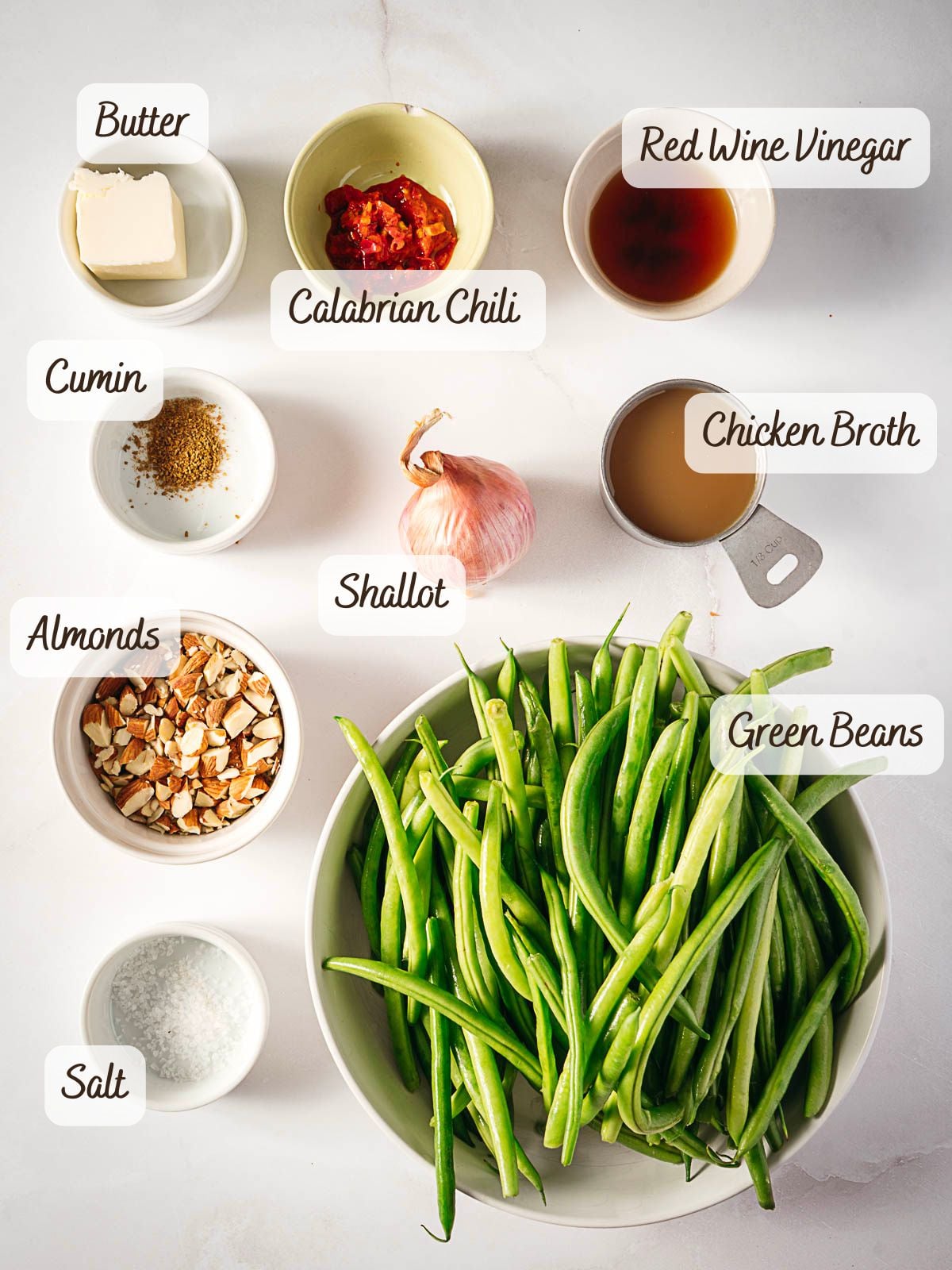 Overhead view of labeled bowls containing butter, Calabrian chili, red wine vinegar, cumin, chicken broth, shallot, green beans, chopped almonds, and salt on a white surface.