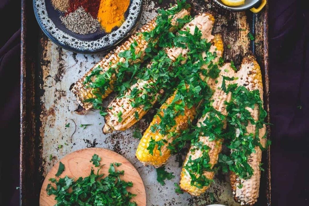 Four grilled corn cobs topped with chopped cilantro on a tray, surrounded by a bowl of spices, lemon wedges, and a small wooden board with extra chopped herbs.