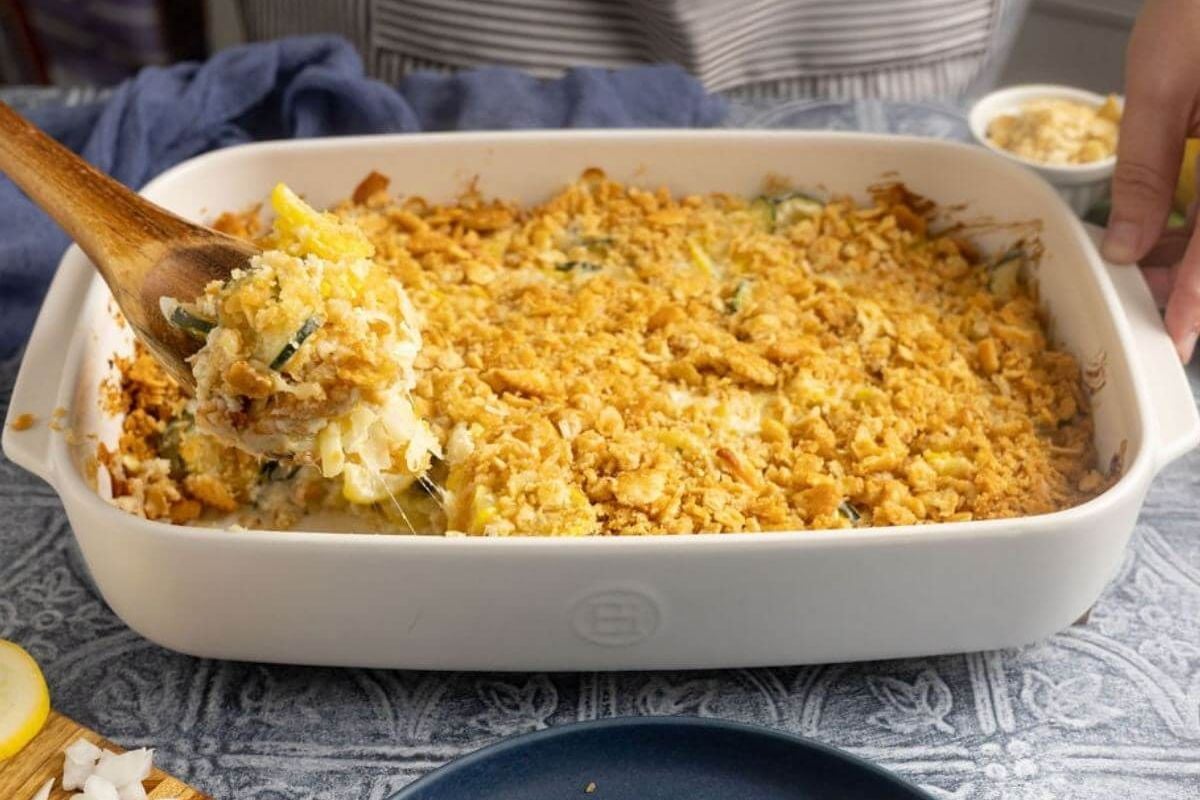 A baked casserole with a golden, crumbly topping is being scooped from a white dish with a wooden spoon.