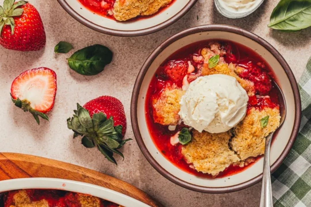 A bowl of strawberry cobbler topped with a scoop of vanilla ice cream. Fresh strawberries and basil leaves are scattered around the table.