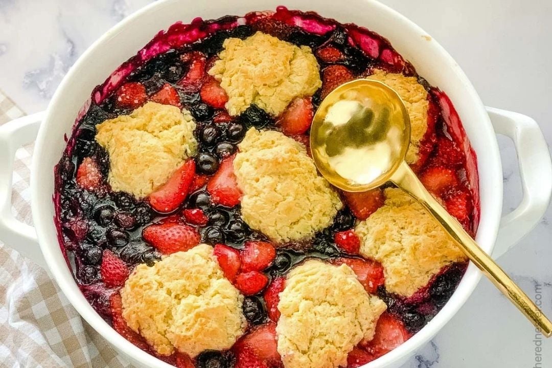 A baked berry cobbler topped with golden biscuit dough sits in a white dish with a gold serving spoon resting on top.