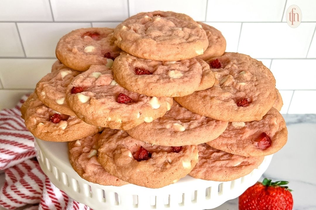 A stack of strawberry cookies with white chocolate chips is displayed on a white cake stand, with a fresh strawberry and red-striped towel nearby.