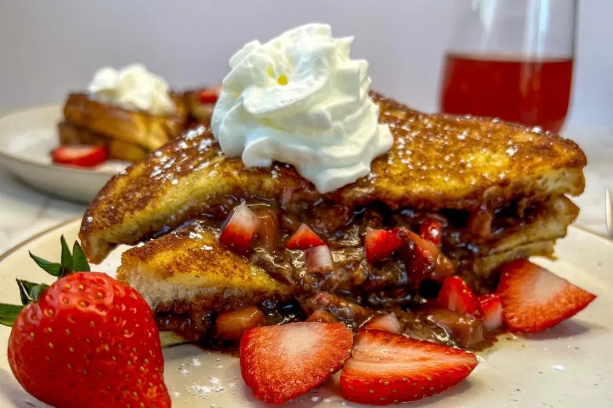 Two slices of stuffed French toast with chocolate and strawberries, topped with whipped cream, are served on a plate with fresh strawberry slices.