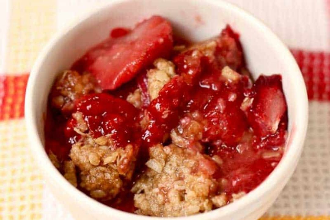 A white bowl filled with strawberry crisp, showing baked oats and sliced strawberries in a red syrup.